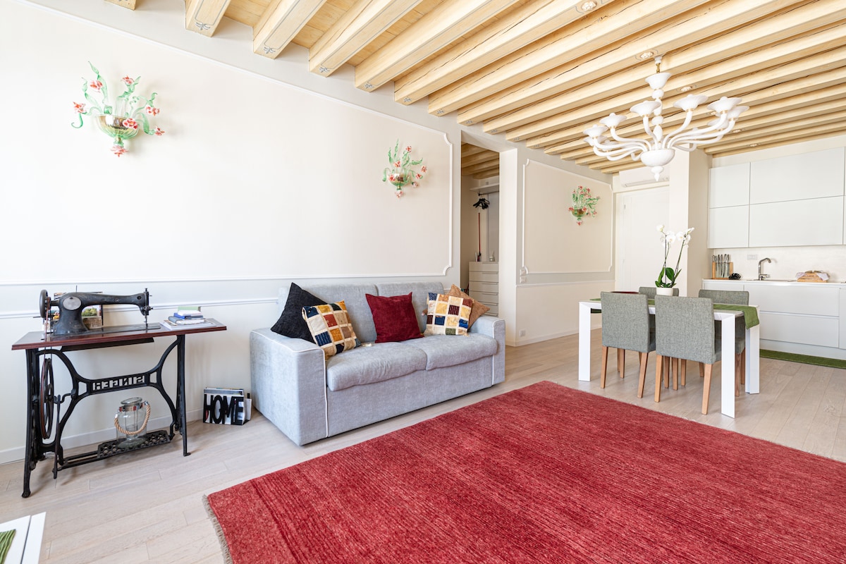 A spacious living area showcases a light-colored sofa adorned with decorative cushions. A vintage sewing machine serves as a side table. A dining table with grey chairs is positioned nearby. The room features a textured red rug and artistic wall sconces providing soft illumination.