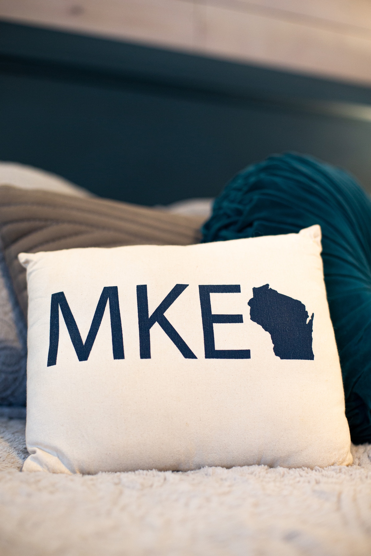 A decorative pillow featuring the letters 'MKE' accompanied by an outline of Wisconsin is positioned on a soft, textured blanket. The pillow, with a neutral background, adds a local touch to the cozy bed setting.