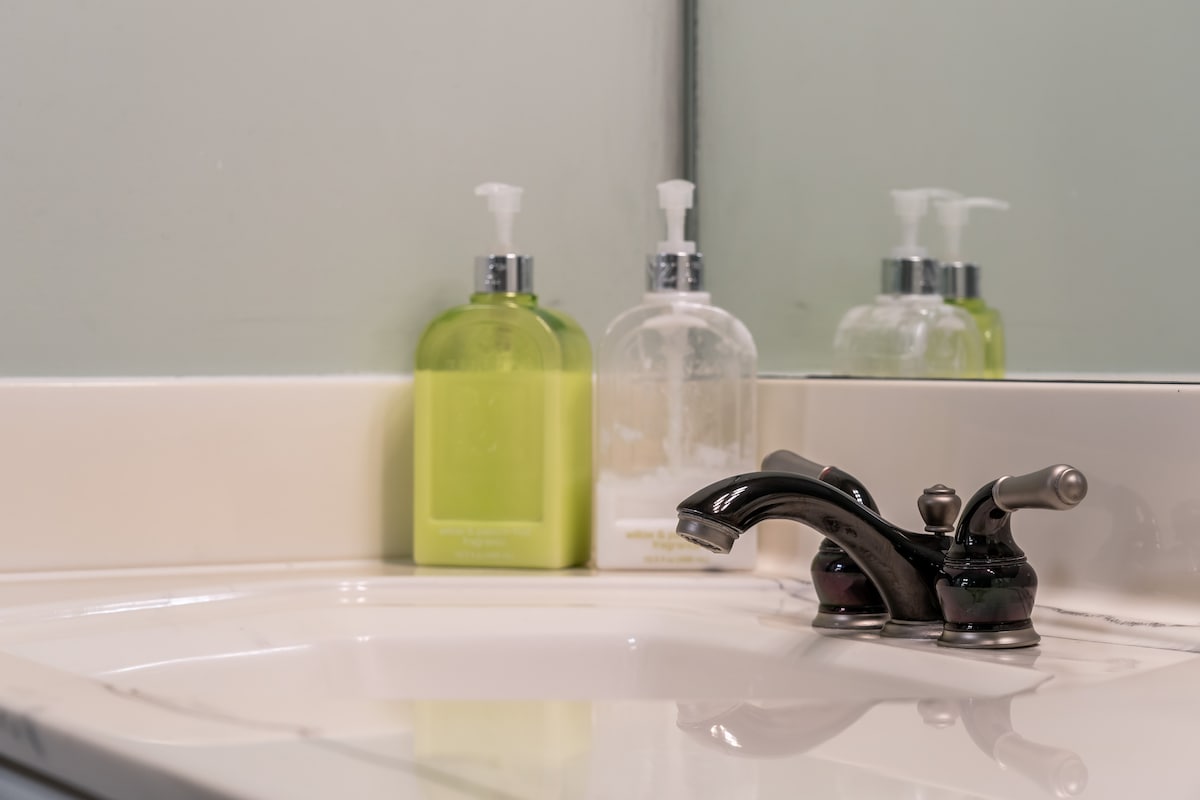 A clean bathroom sink area is featured, showcasing a sleek silver faucet. Two soap dispensers are visible, one with a green label and the other clear, reflecting softly in the mirror above the sink.