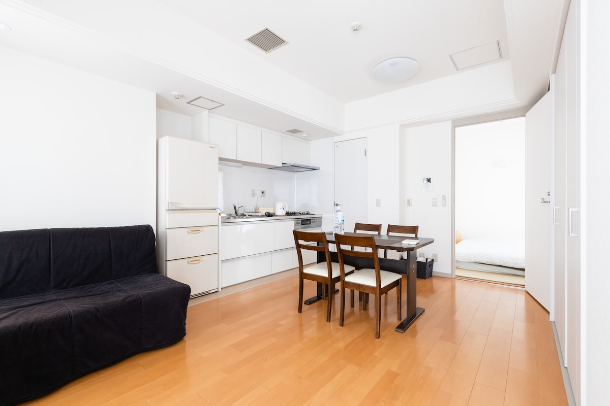 A bright and open living area features a dark sofa and a dining table with four chairs. The kitchen is equipped with modern appliances, and a sliding door leads to a separate sleeping area, enhancing the apartment's functionality.
