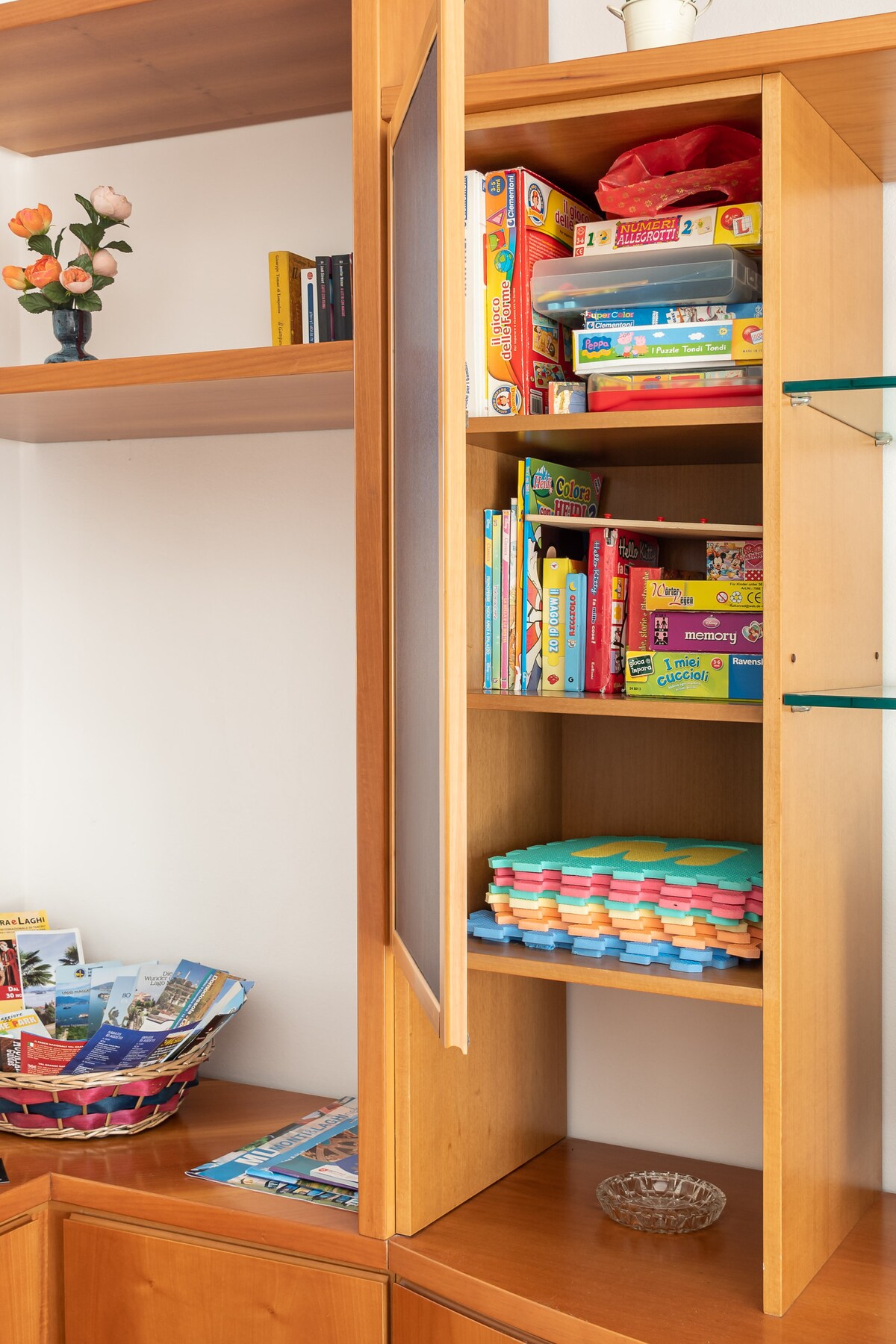 A wooden cabinet with a glass door houses colorful children's books and various games. A basket filled with brochures is placed nearby, alongside a decorative glass dish. A small potted plant adds a touch of greenery to the shelf above.