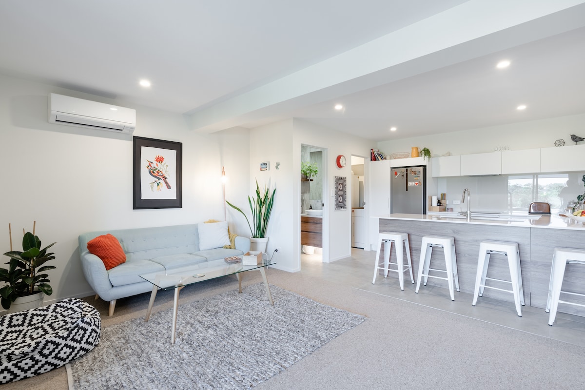 An open living area showcases a light blue sofa paired with a decorative orange pillow. A glass coffee table rests on a textured rug, with a tall plant in the corner. A modern kitchen and dining area can be seen in the background, enhanced by soft lighting.