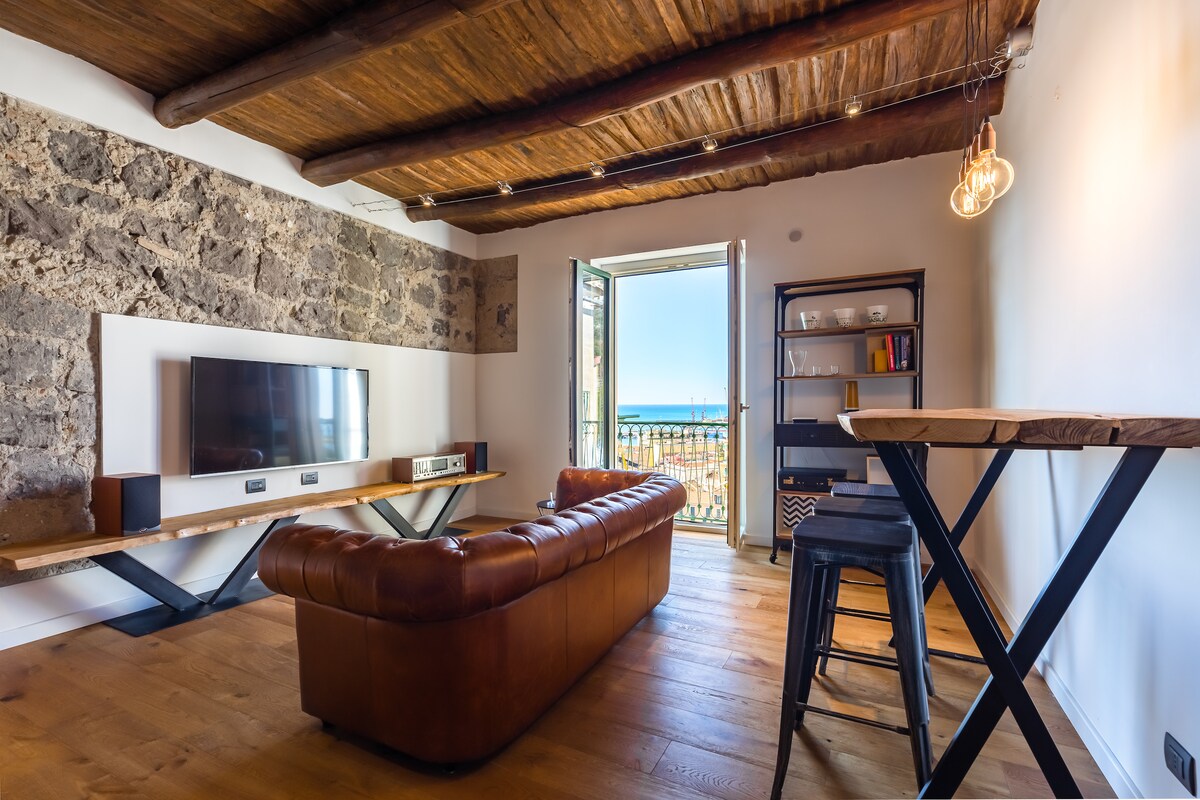 A modern living area features a leather sofa and a sleek wooden table. A flat-screen television is mounted on a textured stone wall. Natural light fills the space through an open door, revealing a sea view and a small balcony beyond.