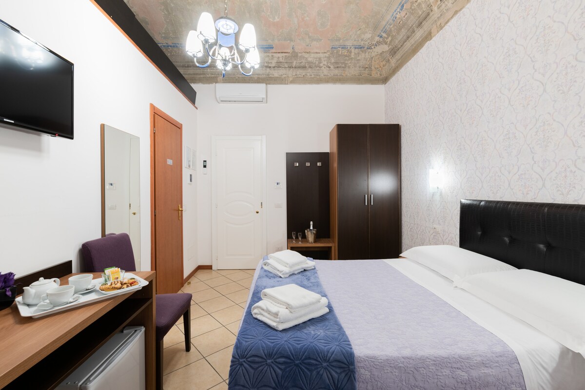 A spacious room is equipped with a comfortable bed dressed in white linens and a soft purple coverlet. A small table displays breakfast items, while a flat-screen TV is mounted on the wall. The room includes a wardrobe and a vintage chandelier overhead.