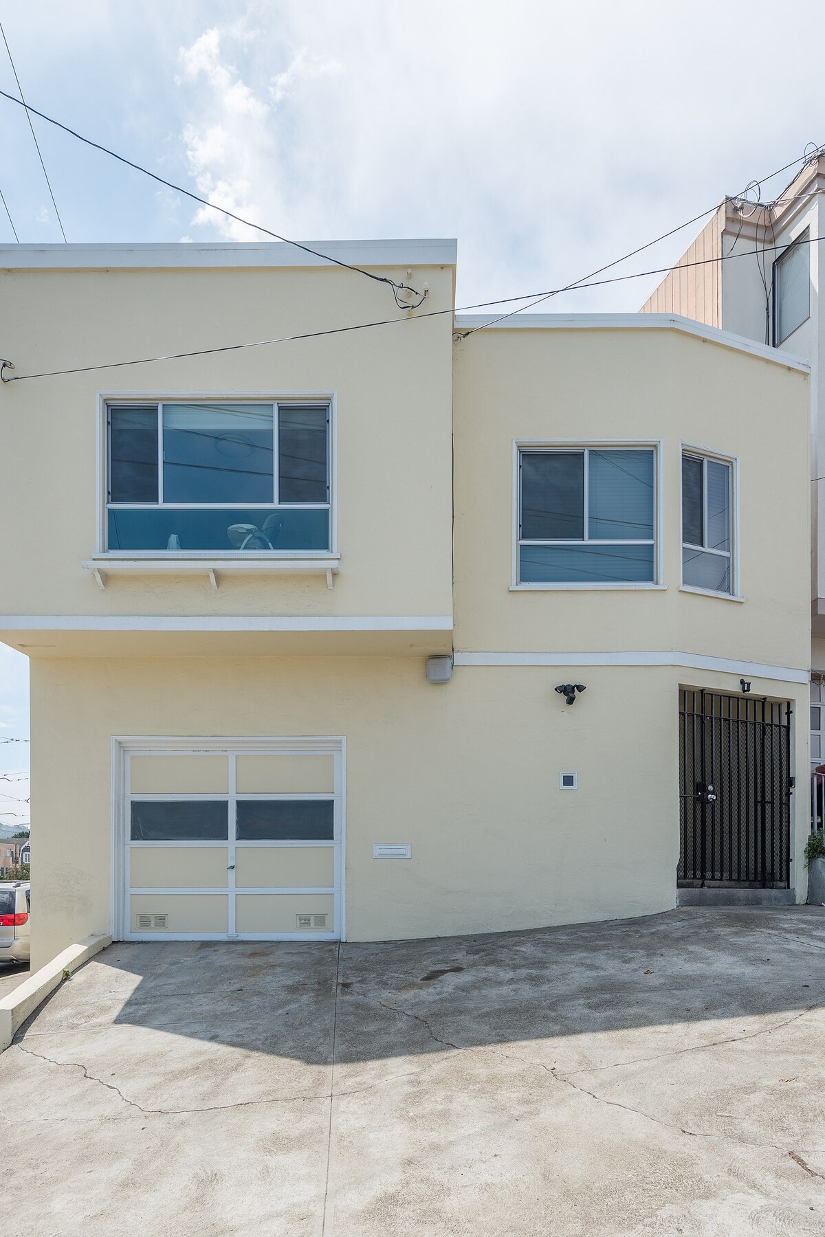An exterior view of the in-law unit is shown, featuring a light yellow facade with multiple large windows. The garage door is visible, accompanied by a gate. The driveway slopes slightly upward, providing access to the entrance.