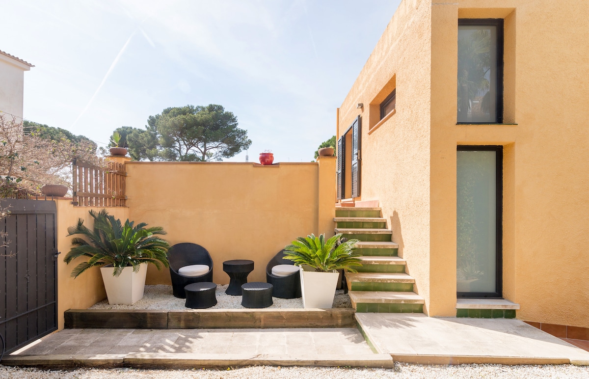 An outdoor seating area is presented, featuring two black chairs and multiple small tables, surrounded by decorative potted plants. A staircase leads to an upper level, while the textured wall offers a warm yellow hue, complementing the natural setting and the clear sky above.