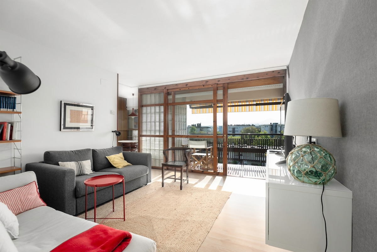 A bright living space is showcased, featuring a gray sofa and a red accent table. Natural light flows through large glass doors opening to a terrace, while a minimalist lamp and decorative elements add to the serene ambiance. A cozy rug enhances the inviting atmosphere.