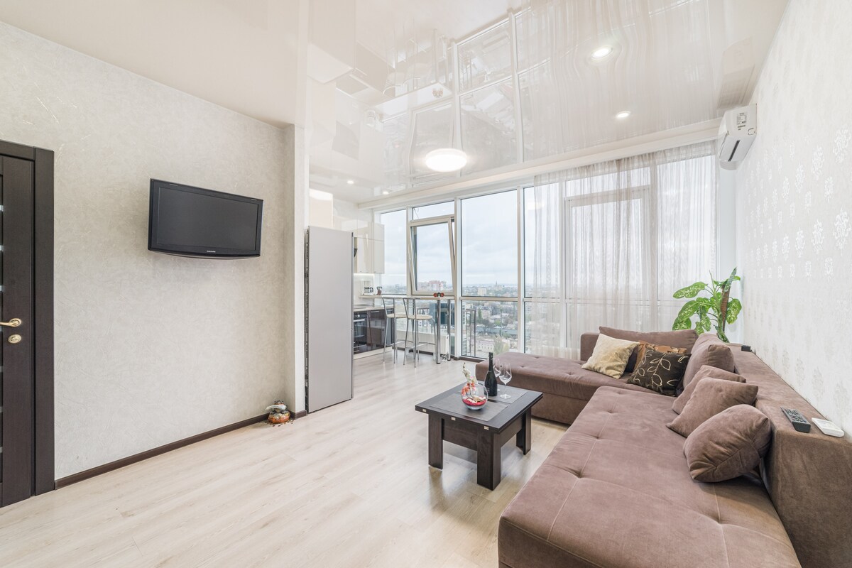 A modern living area features a large brown sectional sofa accented with decorative pillows. A low coffee table is positioned in front, with a television mounted on the wall. Large windows provide natural light, revealing panoramic views of the city and sea.