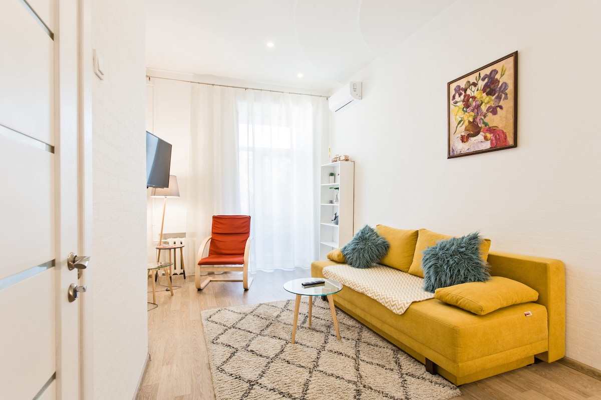 A bright living area is displayed, featuring a yellow sofa with decorative pillows positioned near a coffee table. Natural light enters through sheer curtains, illuminating the space. A television stand and an armchair add to the comfort of this inviting setting.