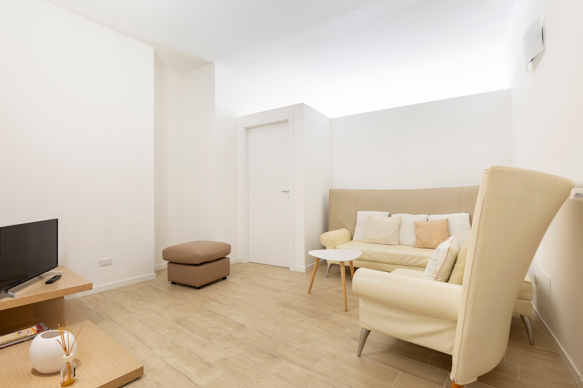The living area features a light-colored couch paired with a matching armchair. A small coffee table rests in the center. A flat-screen television is positioned on a wooden stand. Warm wood flooring extends across the room, complemented by soft lighting from above.