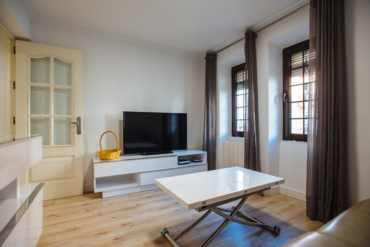 A living area is presented with a modern design, featuring a sleek white coffee table and a flat-screen television mounted on the wall. Natural light enters through two windows, framed by dark curtains, enhancing the simplicity of the light-colored walls and wooden flooring.