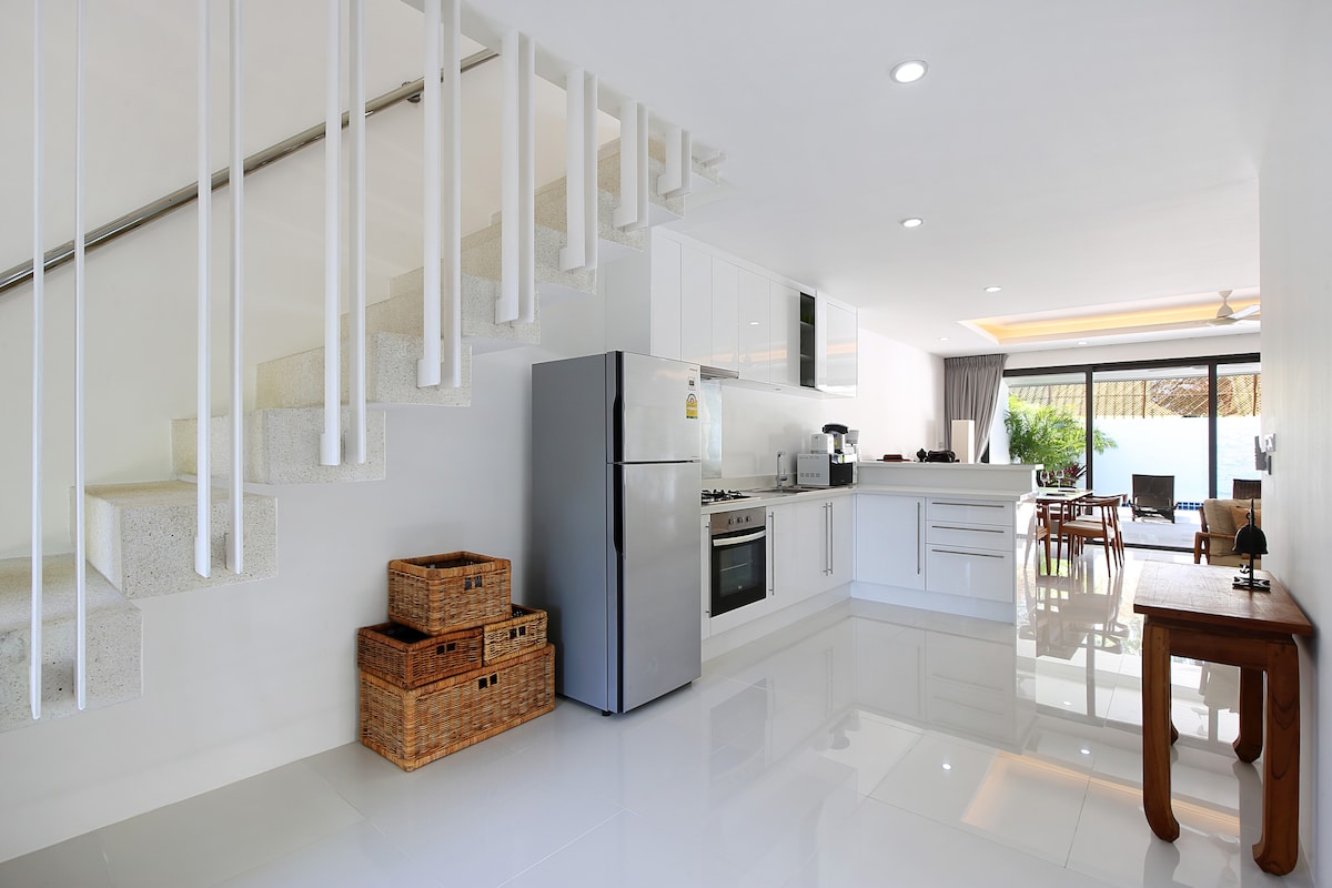 A modern open-plan kitchen and dining area is visible, featuring sleek white cabinetry and stainless steel appliances. Natural light floods the space through large windows, illuminating the polished floor. A staircase with a minimalist design leads to the upper level, while wicker storage baskets add functionality.