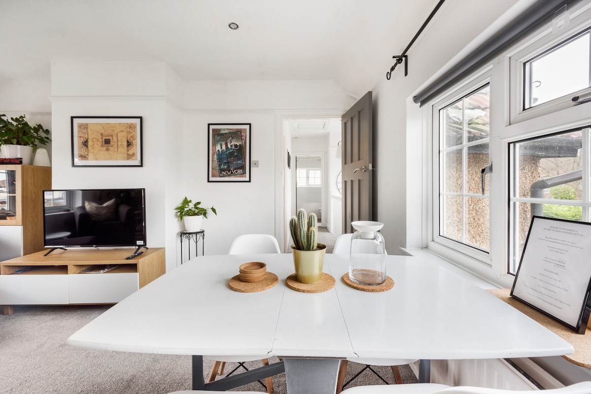 A bright dining area features a white table surrounded by modern chairs. A potted cactus stands on the table alongside a glass jar. A television is positioned on a low wooden cabinet against the wall, with subtle decor and natural light coming through the window.