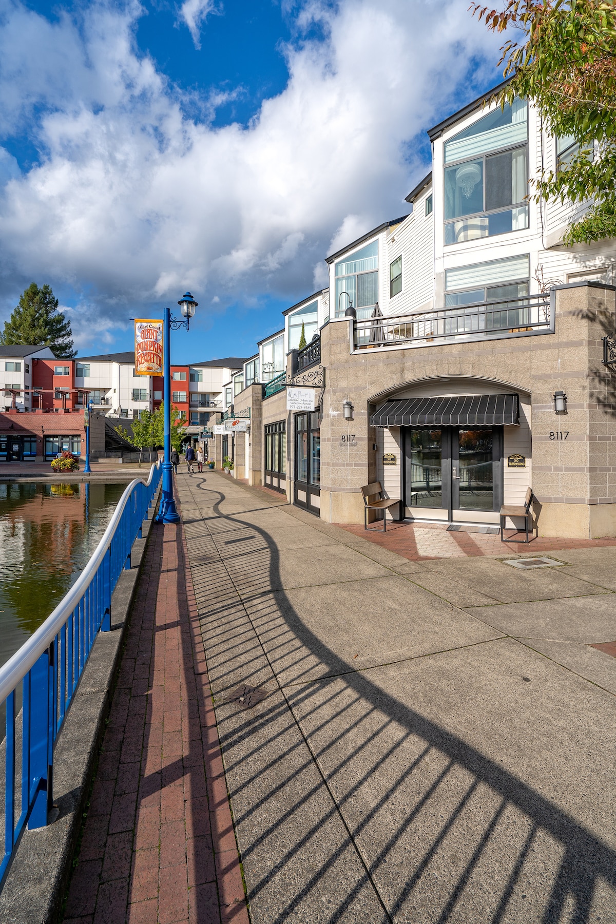 The image depicts a charming walkway alongside a man-made lake, flanked by modern condo units. A decorative blue railing outlines the path, which features both concrete and brick paving. The bright sky is accentuated by scattered clouds, creating a vivid backdrop for the lakeside setting.