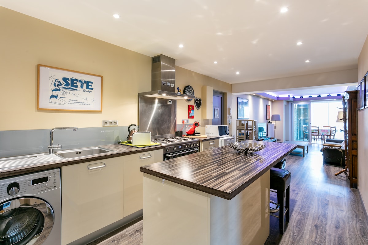 A modern kitchen features a spacious island with stools and is equipped with a washing machine, oven, and other appliances. Light-colored cabinetry and wooden accents create a warm atmosphere, while a dining area is visible in the background, illuminated by natural light from large windows.