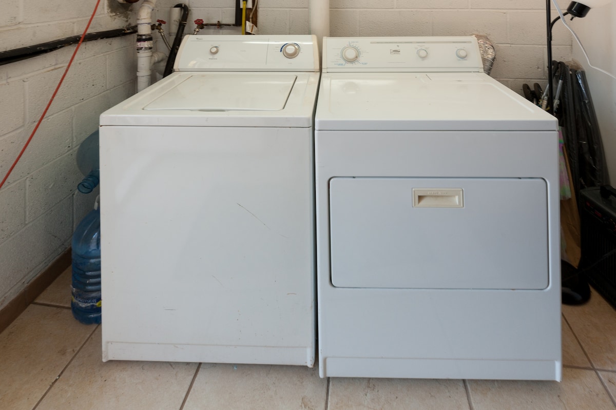 A washing machine and a dryer are positioned side by side, featuring white exteriors with minimalist designs. The washing machine has a top-loading door, while the dryer showcases a front-loading compartment. The appliances sit on tiled flooring, with plumbing visible in the background.