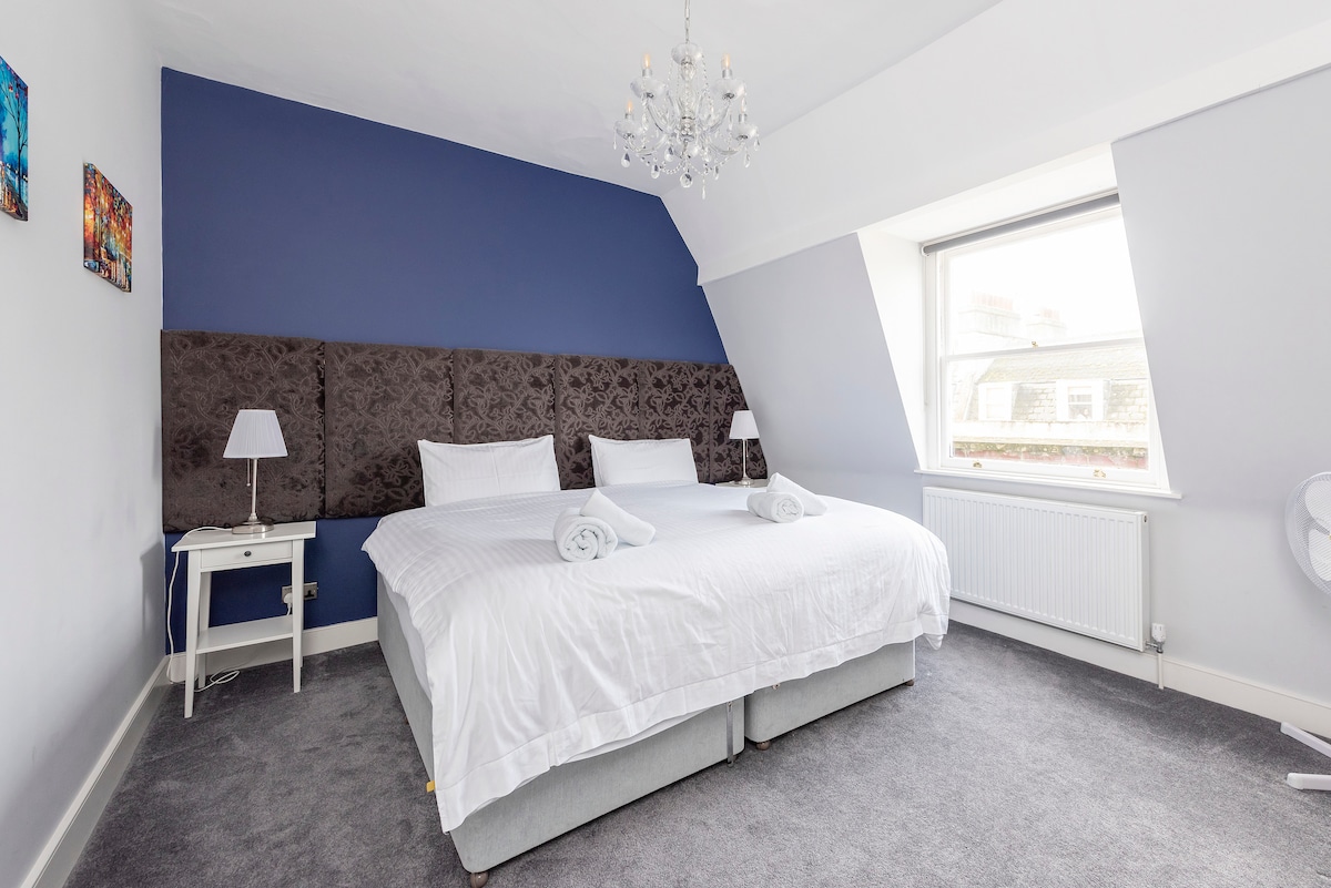 A spacious bedroom features a large bed with plush bedding, complemented by bedside lamps on either side. The wall behind the bed is adorned with a deep blue hue, and bright artwork adds a touch of color. A large window offers natural light and views outside.
