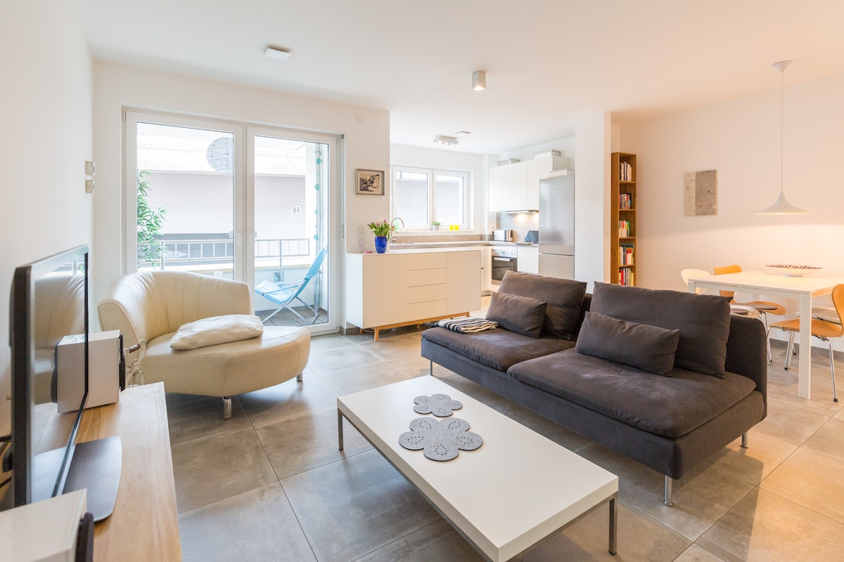 A modern living area features a plush gray sofa and a light-colored coffee table. Large glass doors open to an outdoor space, while a well-equipped kitchen is visible in the background. The dining area includes a round table with four chairs. Natural light fills the room.