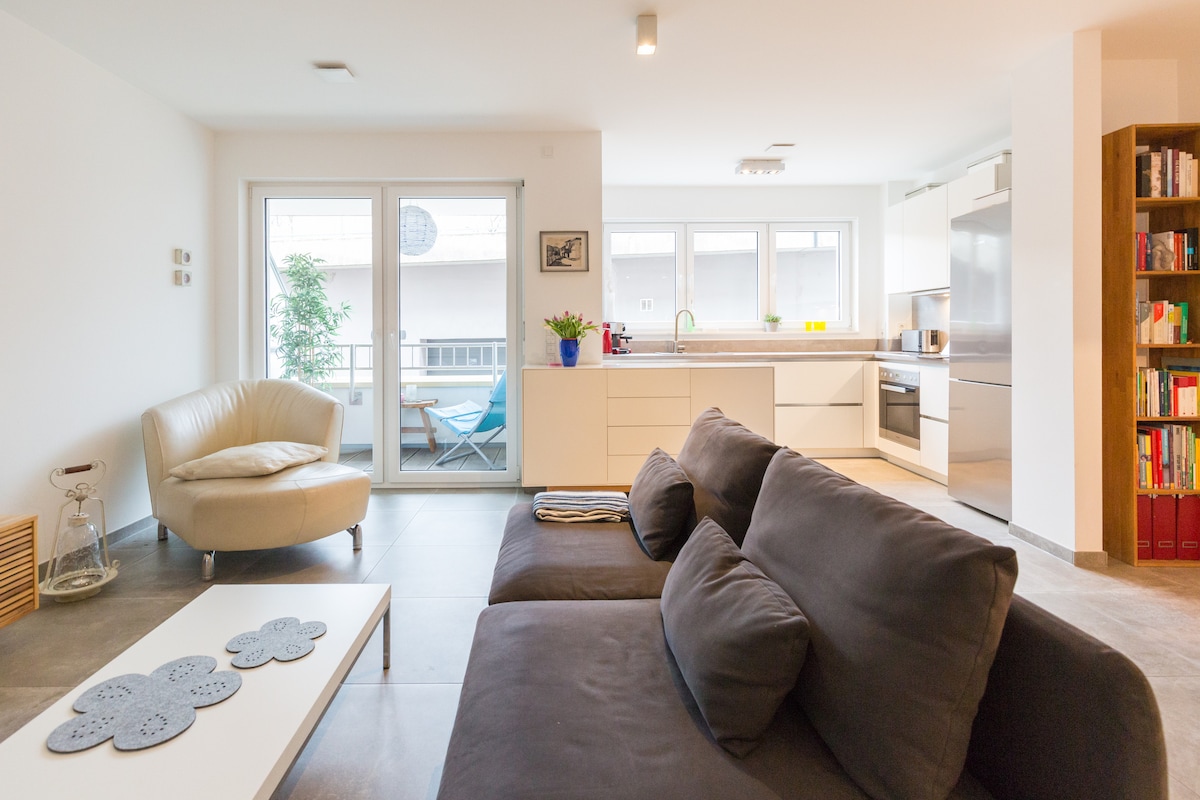 The living area features a comfortable dark gray sofa and a light-colored coffee table, accented by a decorative placemat. A modern kitchen with sleek cabinetry is visible, and large windows allow natural light to brighten the space, which opens to a small balcony with outdoor seating.