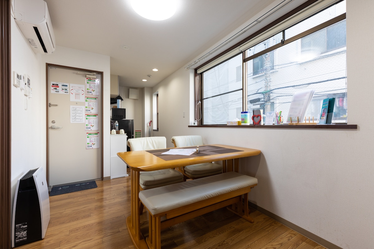 A dining area is featured with a minimalist wooden table and light-colored benches. Natural light streams in through a large window, illuminating the space. A compact kitchenette is visible in the background, along with various informational materials displayed on the wall.