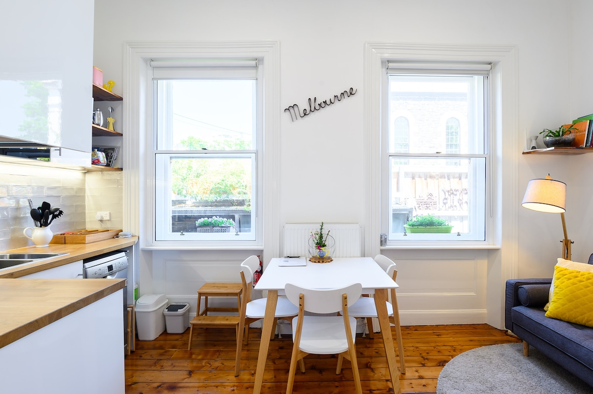 The kitchen and living area feature a light-filled space with large windows, allowing natural light to illuminate the room. A dining table with four chairs is positioned beside the windows, while a cozy sofa is placed across from the table. Decorative elements include the word 'Melbourne' on the wall.