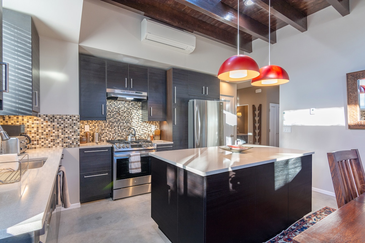The modern kitchen features dark cabinetry and stainless steel appliances, including a gas stove. A large island with a light-colored countertop provides space for preparation and dining. Two red pendant lights hang above the island, and natural light brightens the room.