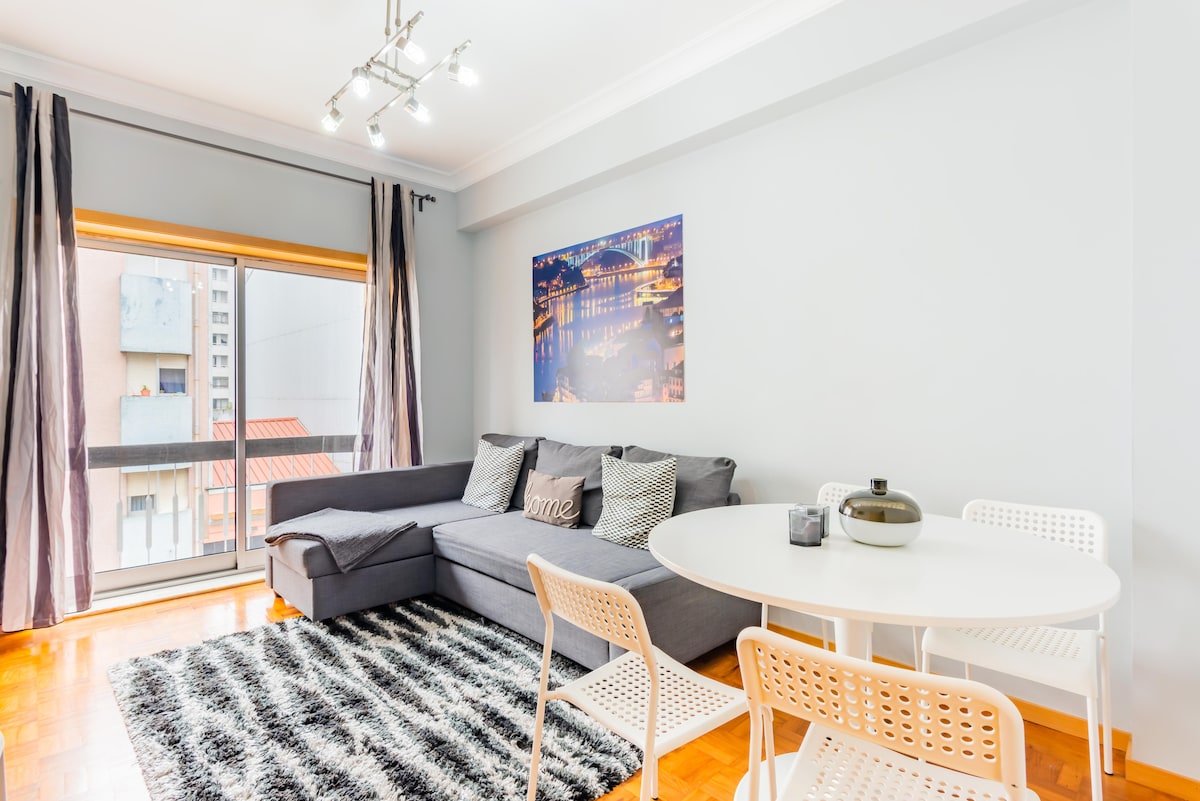 A modern living area features a comfortable sectional sofa with decorative cushions and a soft area rug. A circular dining table is surrounded by four white chairs. Natural light enters through a balcony door, which is flanked by dark curtains, enhancing the bright and airy space.