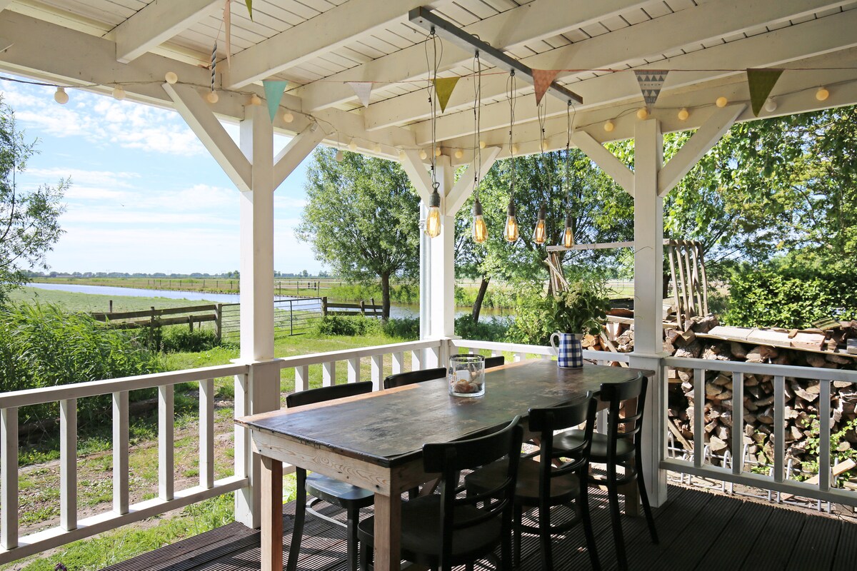 A large porch is featured, offering an outdoor dining area with a spacious wooden table and several chairs. Decorative lights hang above, illuminating the setting. The view showcases meadows and greenery, creating a serene and open atmosphere.