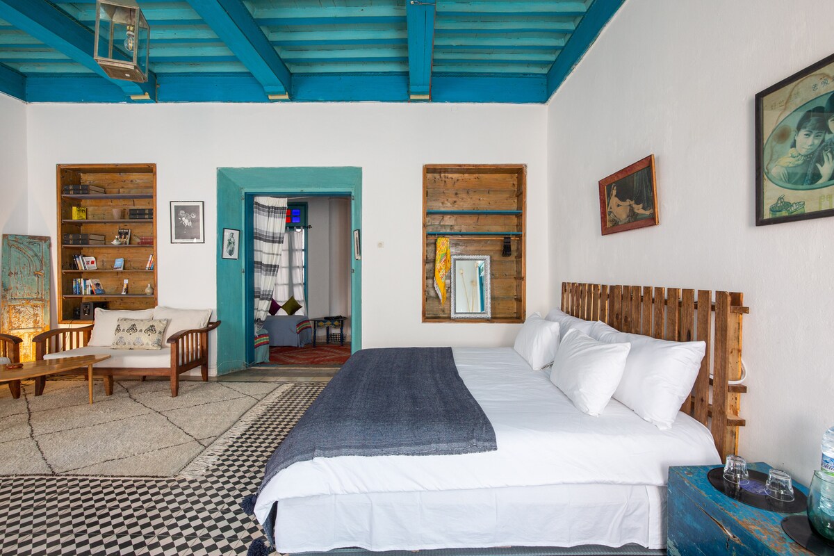 A spacious bedroom features a comfortable bed with plush white pillows and a textured navy blanket. Bright blue wooden beams contrast with traditional zellige flooring. Shelves adorned with decor and a seating area are visible, adding to the room's airy feel.