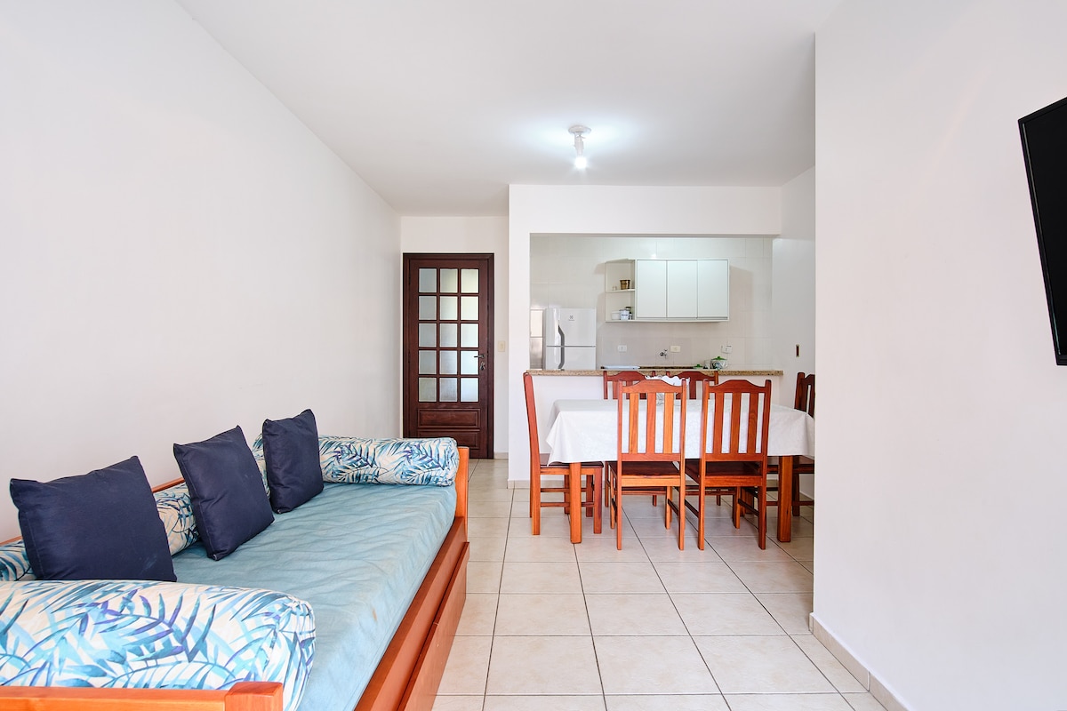 An open living area is presented with a light-colored tiled floor. A blue sofa with tropical-themed cushions is positioned beside a dining table with wooden chairs. A kitchenette is visible in the background, equipped with modern appliances and bright, neutral cabinetry.