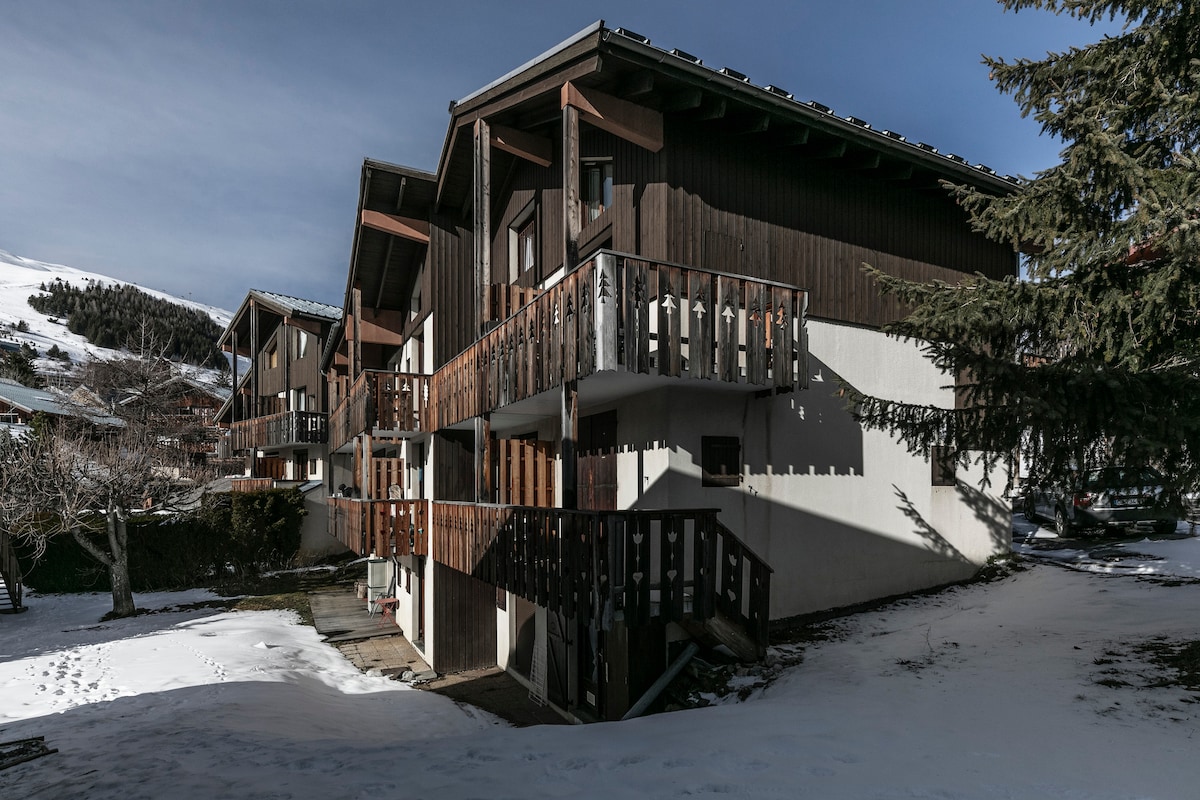 The exterior of a multi-unit building is depicted, featuring wooden balconies and a mix of dark wood and white siding. Snow covers the ground, while the surrounding landscape is partially visible. Tall trees are present nearby, contributing to the serene setting.