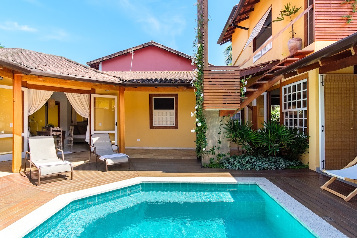An inviting outdoor area features a sparkling pool surrounded by a wooden deck. Several lounge chairs are positioned nearby, and lush greenery adds to the serene atmosphere. The living area is visible through open doors, showcasing a warm and welcoming space.