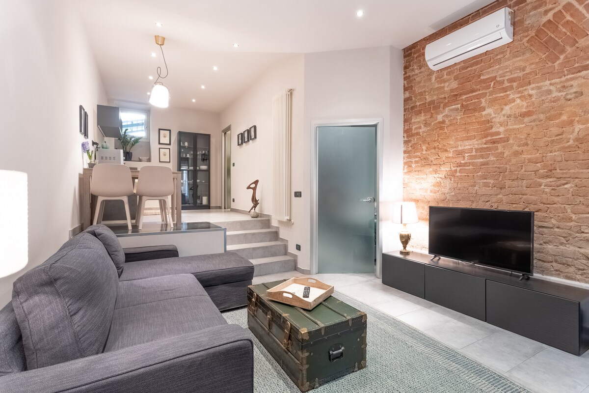 The living area features a comfortable gray sofa and a wooden coffee table, complemented by a stylish rug. An exposed brick wall adds texture, while a 40" smart TV is positioned on a modern black cabinet. Natural light illuminates the space, enhancing the inviting atmosphere.