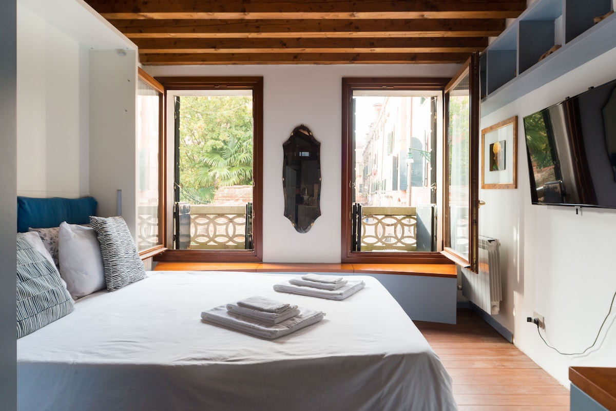 A bright sleeping area features large windows providing views of a serene Venetian courtyard. A spacious bed is dressed in white linens, complemented by neatly arranged towels. Exposed wooden beams enhance the architectural character of the room, while a flat-screen TV is mounted on the wall.