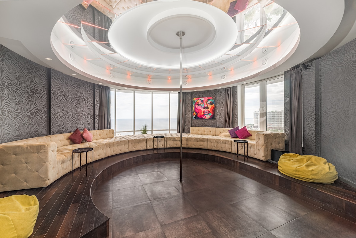 A spacious lounge area features a circular arrangement of plush seating with decorative cushions. Large windows provide expansive views of the sea, while a sleek pole is centrally positioned. The elegant ceiling design complements the inviting atmosphere, and soft lighting enhances the overall space.