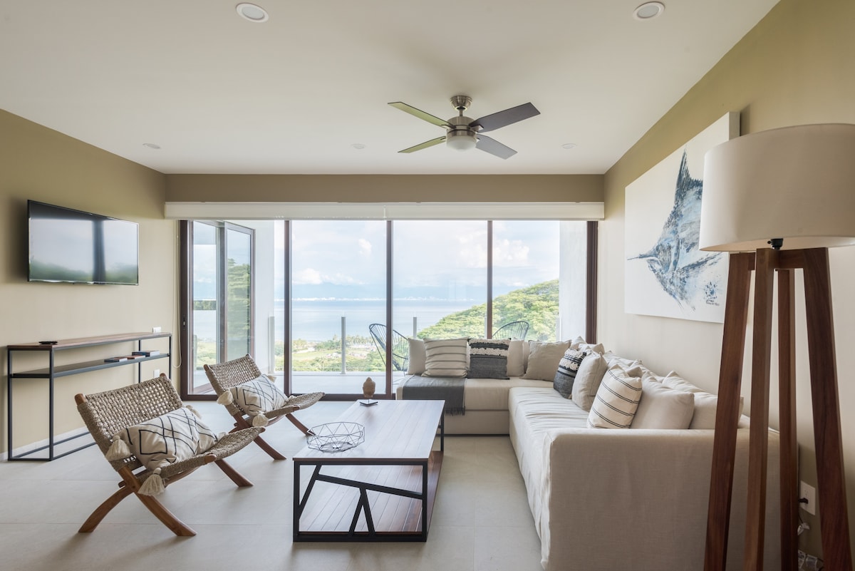 A bright and airy living area is showcased, featuring large glass sliding doors that offer unobstructed ocean views. A sectional sofa is accompanied by a wooden coffee table, and two woven chairs add texture. A ceiling fan and wall-mounted TV enhance the comfort of this inviting space.