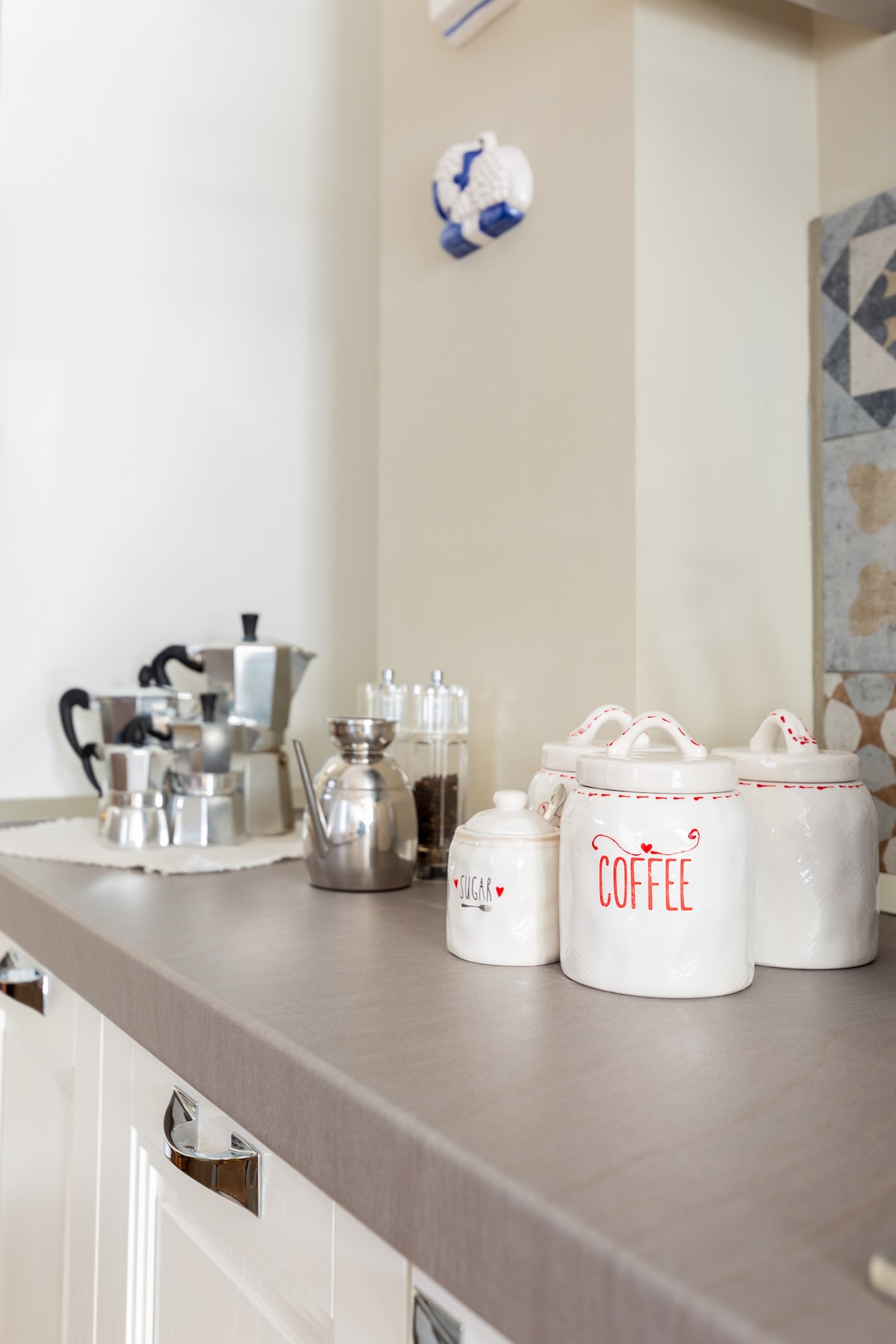 A neatly arranged kitchen countertop is visible, featuring various coffee-making equipment. Included are metal coffee pots, glass containers, and white canisters labeled 'COFFEE.' The surface is light gray, providing a clean and organized appearance.