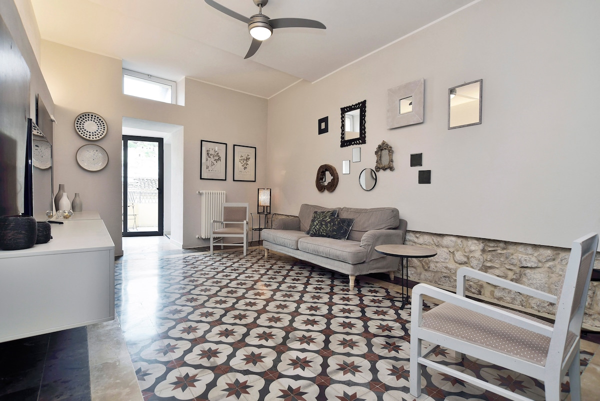 A spacious living area features a comfortable gray sofa and a white armchair, arranged on intricately designed tiled flooring. Wall decorations, including mirrors and framed art, add charm. Natural light enters through a nearby window, complementing the neutral color palette of the room.