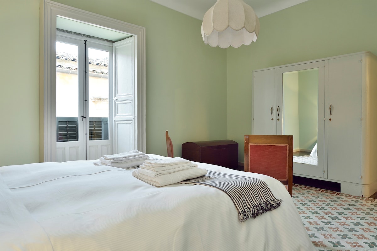 A serene bedroom features a spacious bed with neatly folded linens. Light green walls complement the patterned tiled floor. A wardrobe with mirrored doors stands opposite a wooden chair, while natural light filters in through open double doors leading to a small balcony.