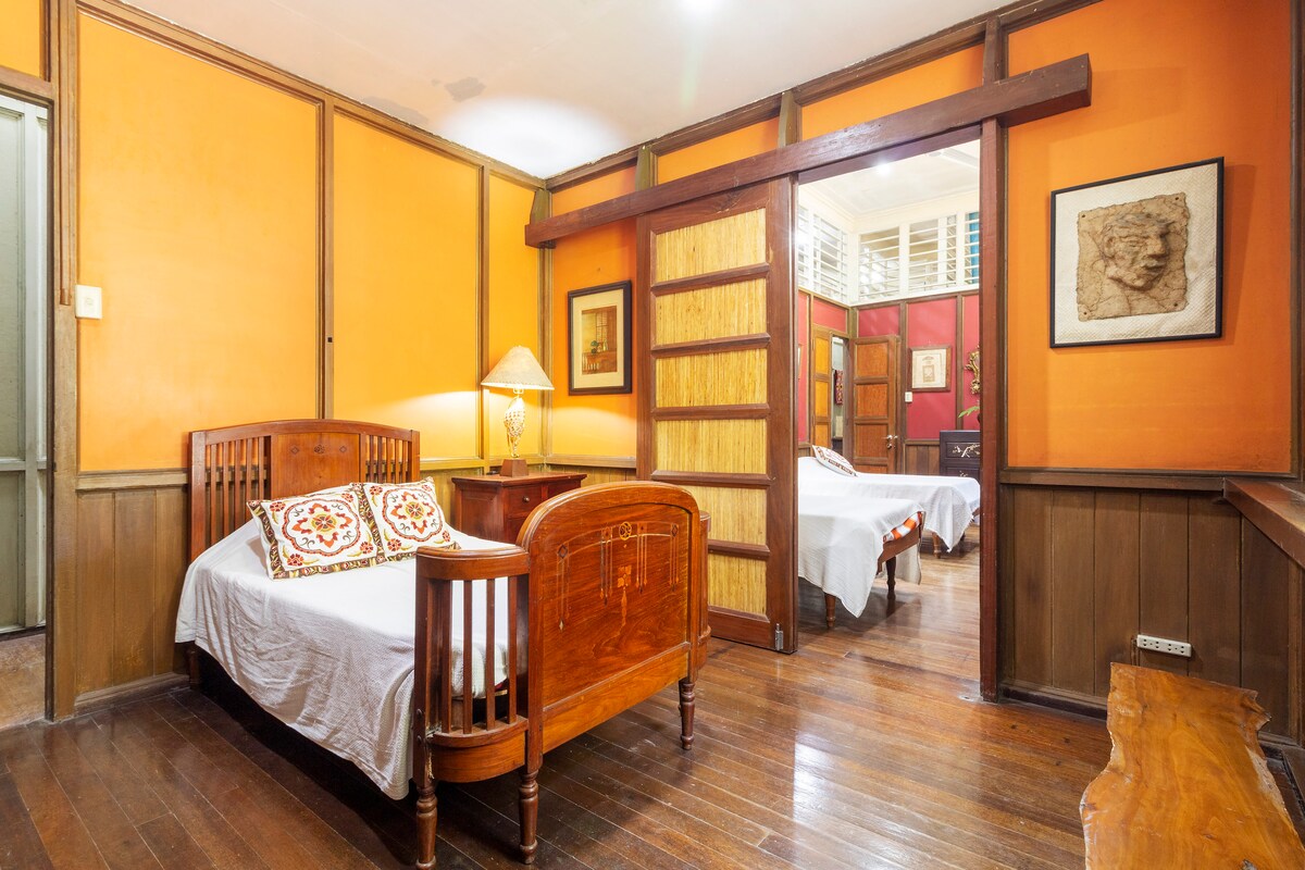 A guest room features warm orange walls and polished wooden floors. Two wooden beds are positioned across from each other, each adorned with white linens and decorative pillows. A standing lamp provides soft lighting, while art pieces and sliding wooden doors enhance the room's character.