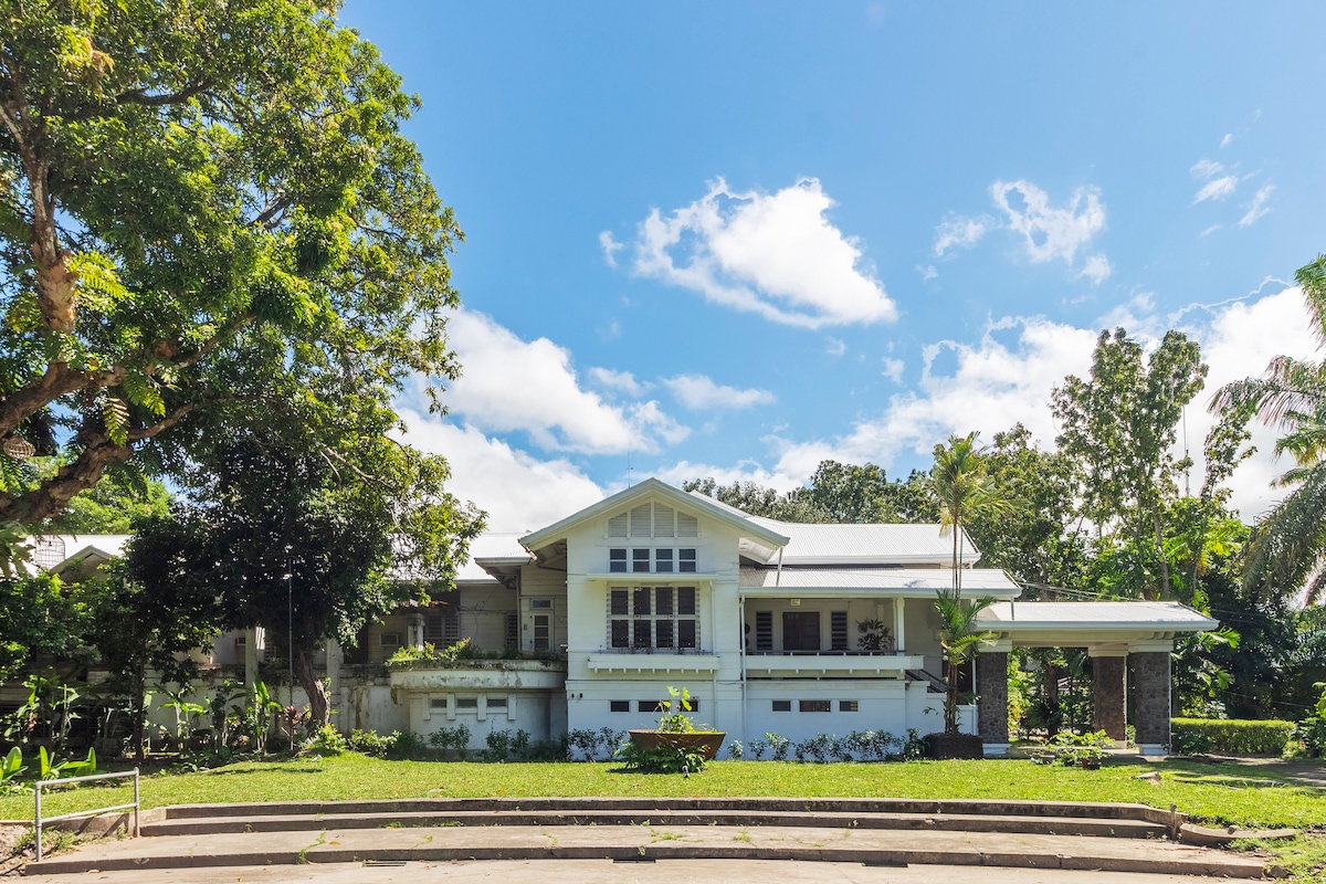 A spacious house is set within a lush green landscape, showcasing a large front porch and multiple windows that invite natural light. The structure is surrounded by trees and tropical foliage, presenting a tranquil environment. A clear blue sky provides a serene backdrop.