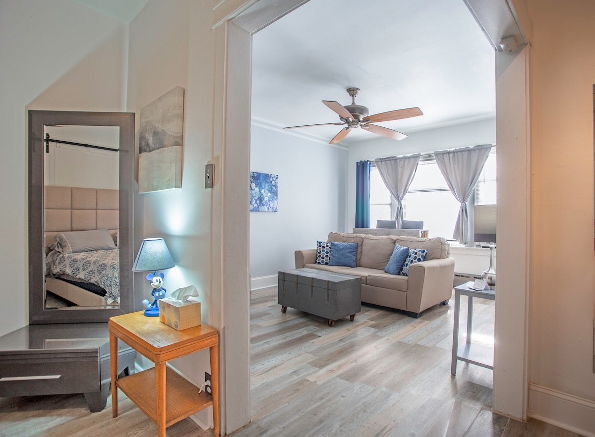 A bright living space featuring a beige sofa adorned with decorative pillows, adjacent to a coffee table and a small side table with a lamp. A ceiling fan provides airflow, and large windows dressed with sheer curtains allow natural light to fill the area. A bedroom area is visible through an open doorway.