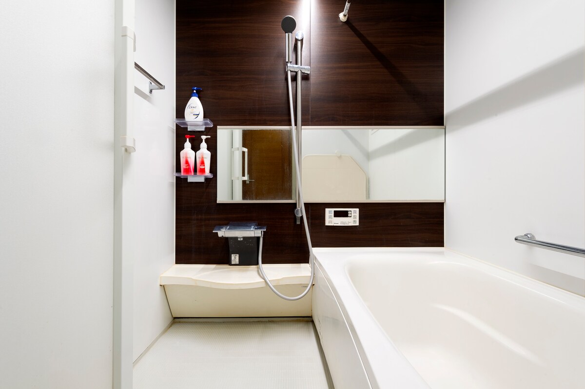 A contemporary bathroom features a bathtub with a sleek, curved design. The dark wood accent wall is complemented by a full-length mirror and a showerhead mounted on the wall. Bottles of toiletries are neatly arranged on a shelf within easy reach.