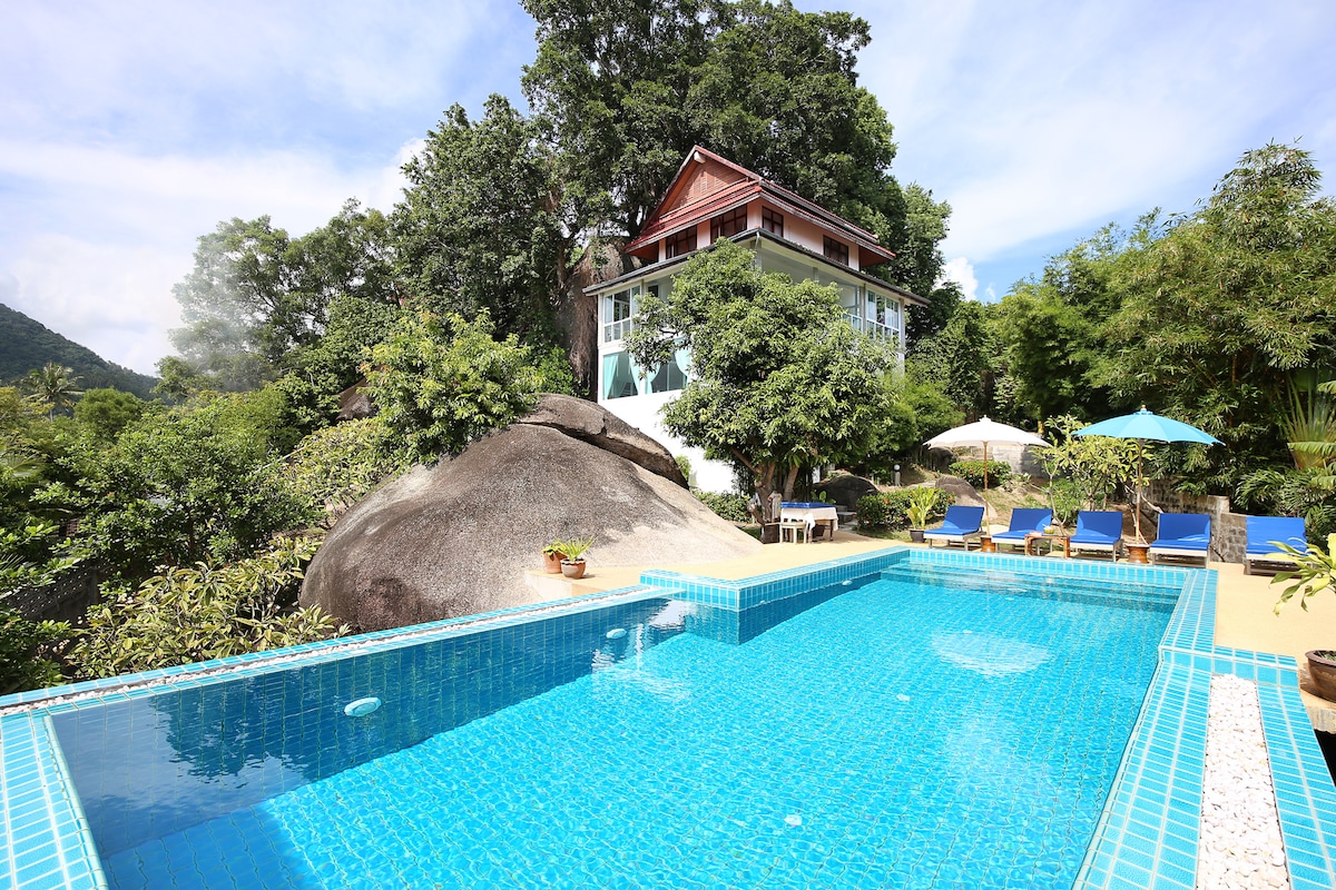 A swimming pool is set in the foreground, surrounded by lush greenery and sun loungers. The villa is visible in the background, standing amidst large rocks and trees. Umbrellas provide shaded seating areas, and the vibrant blue water complements the natural landscape.
