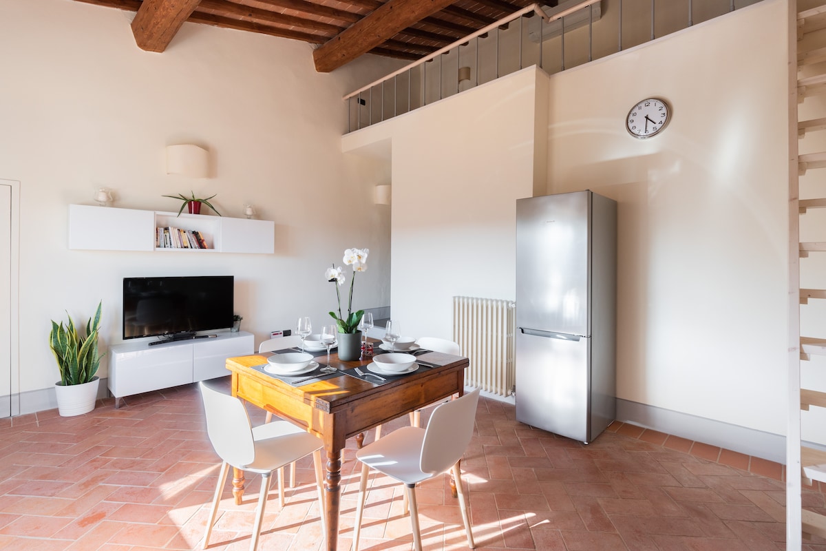 A dining area is depicted, featuring a wooden table set for four, complemented by white chairs. A modern stainless-steel refrigerator and a flat-screen television are visible along the wall, with a decorative plant adding a touch of greenery.