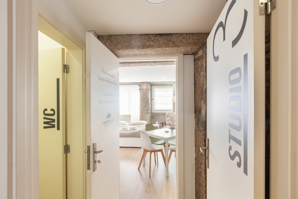 The image shows an entryway with two doors labeled 'STUDIO' and 'WC'. A glimpse of the open space can be seen through the doorway, revealing modern furniture and natural light illuminating the area. The walls are adorned with a mix of granite and contemporary finishes.