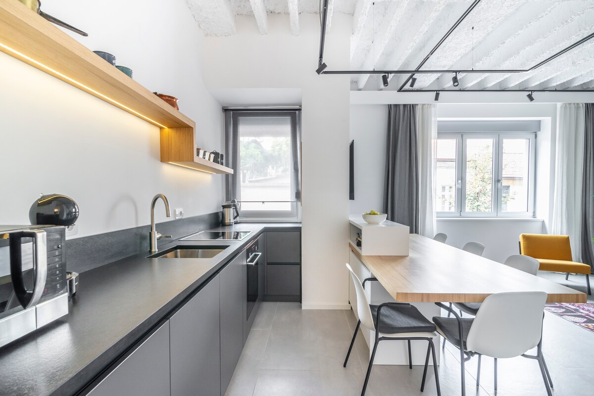 A modern kitchen showcases sleek cabinetry and a spacious countertop. A dining area features a large table with six chairs. Natural light floods the space through a window, complementing the minimalist design of the gray and wood elements.