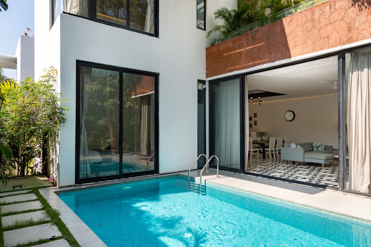 A private pool is framed by a stylish modern villa, featuring large glass doors that open to a spacious living area. Lush greenery surrounds the property, enhancing the serene outdoor environment. The pool is designed for relaxation, reflecting the inviting atmosphere of the villa.