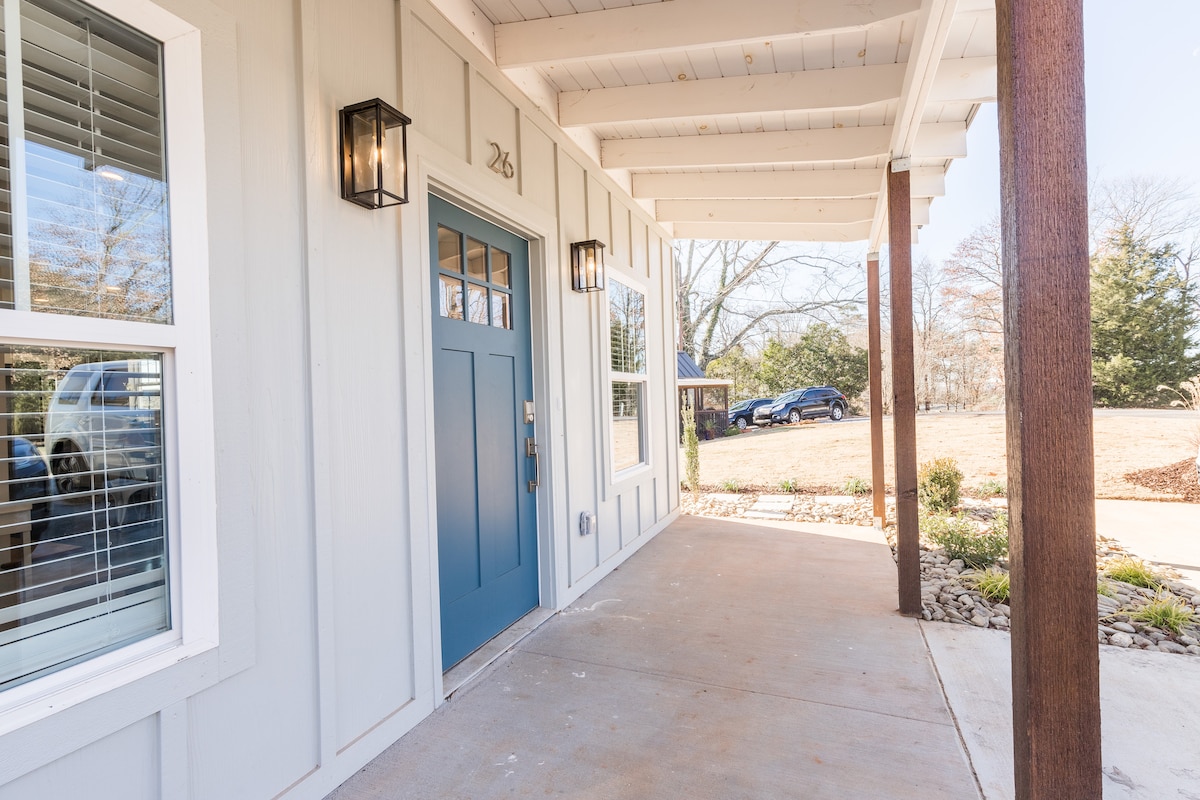 A welcoming entrance is displayed with a blue front door framed by two lanterns. The covered porch features a smooth concrete floor and is surrounded by light-colored siding. A grassy area is visible in the background, alongside a gravel pathway and a parked car.