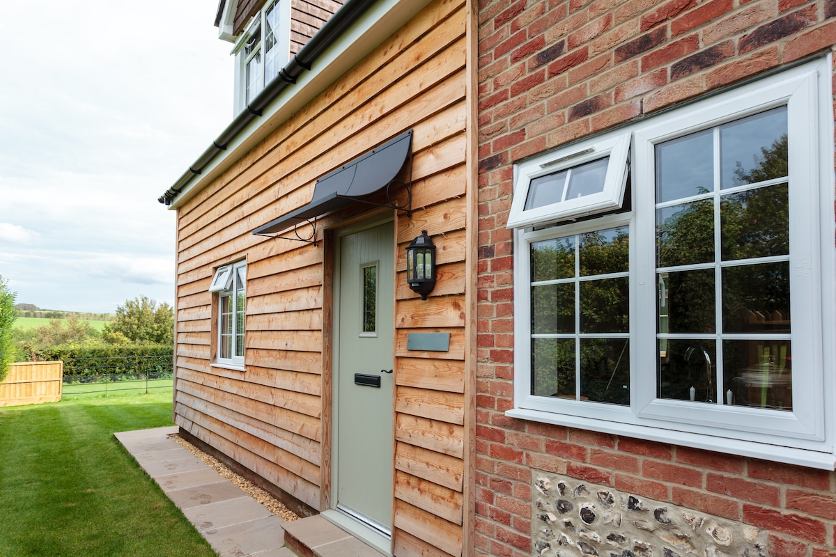 The exterior of the cottage is characterized by a combination of warm, wooden cladding and red brick. A welcoming front door is complemented by a lantern fixture and decorative awning. Nearby, multiple window panes reflect the surrounding greenery.